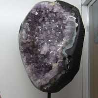 Amethyst crystal formation on a black stand against a white wall.