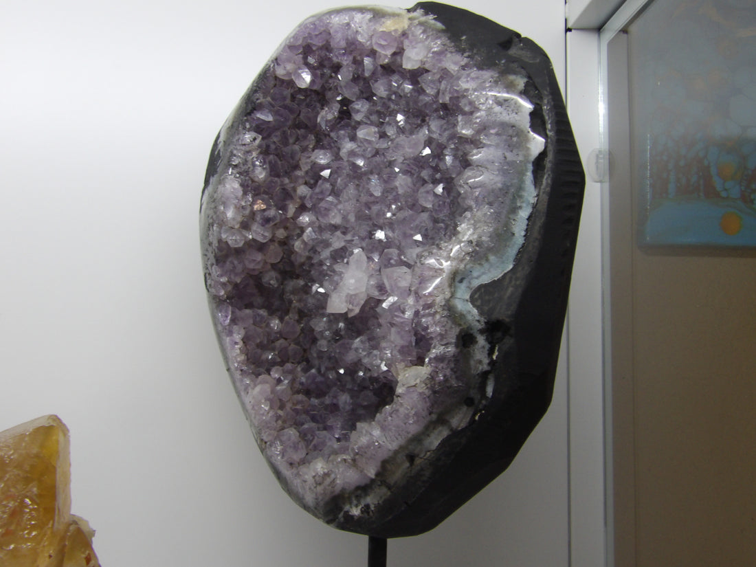 Amethyst crystal formation on a black stand against a white wall.